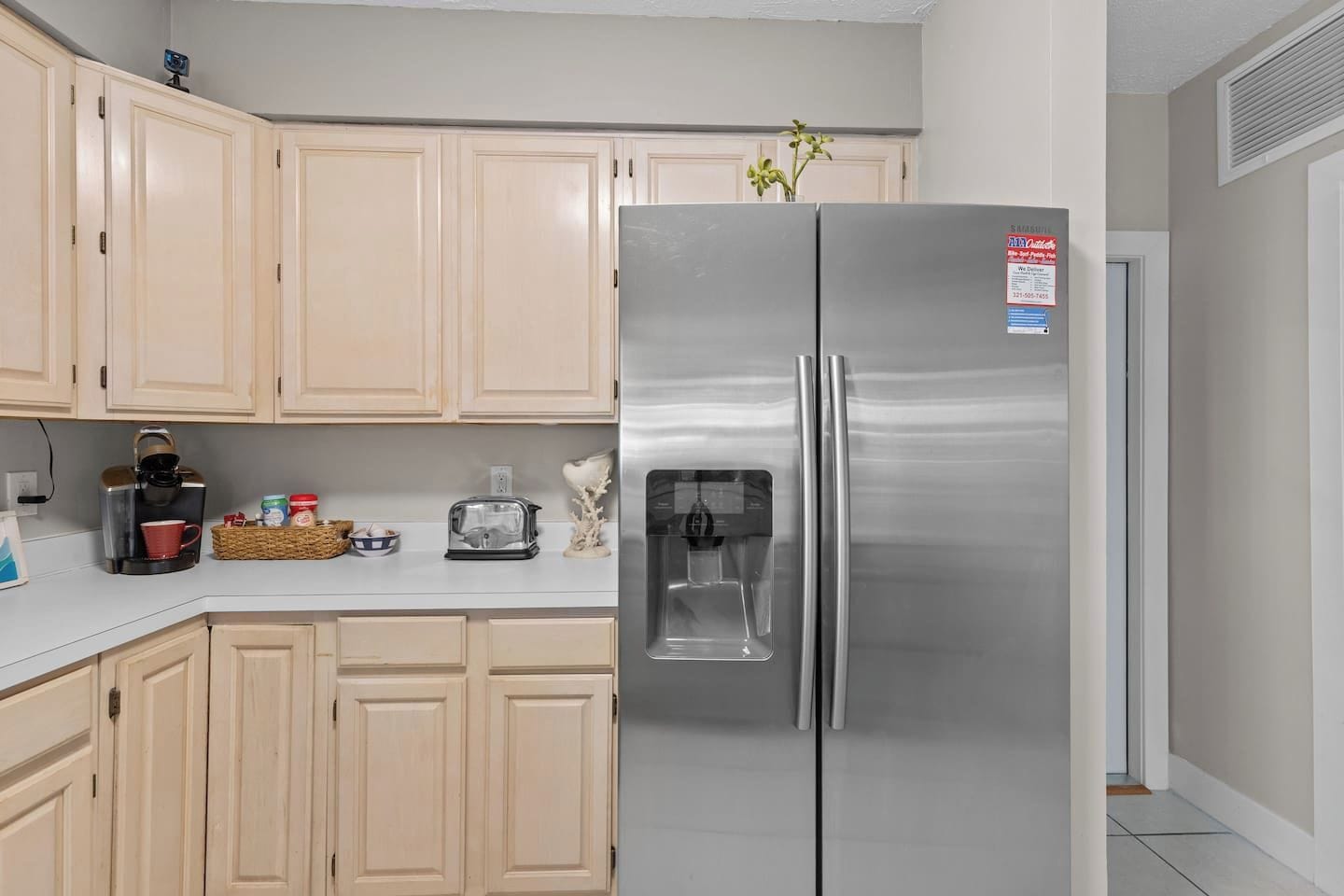 Una cocina con armarios de colores claros, un refrigerador de acero inoxidable, cafetera, tostadora y una pequeña planta.