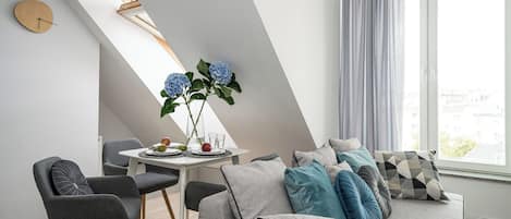 A modern living room with a dining area, featuring a sofa set, a dining table with chairs, a clock on the wall, and a window with curtains.
