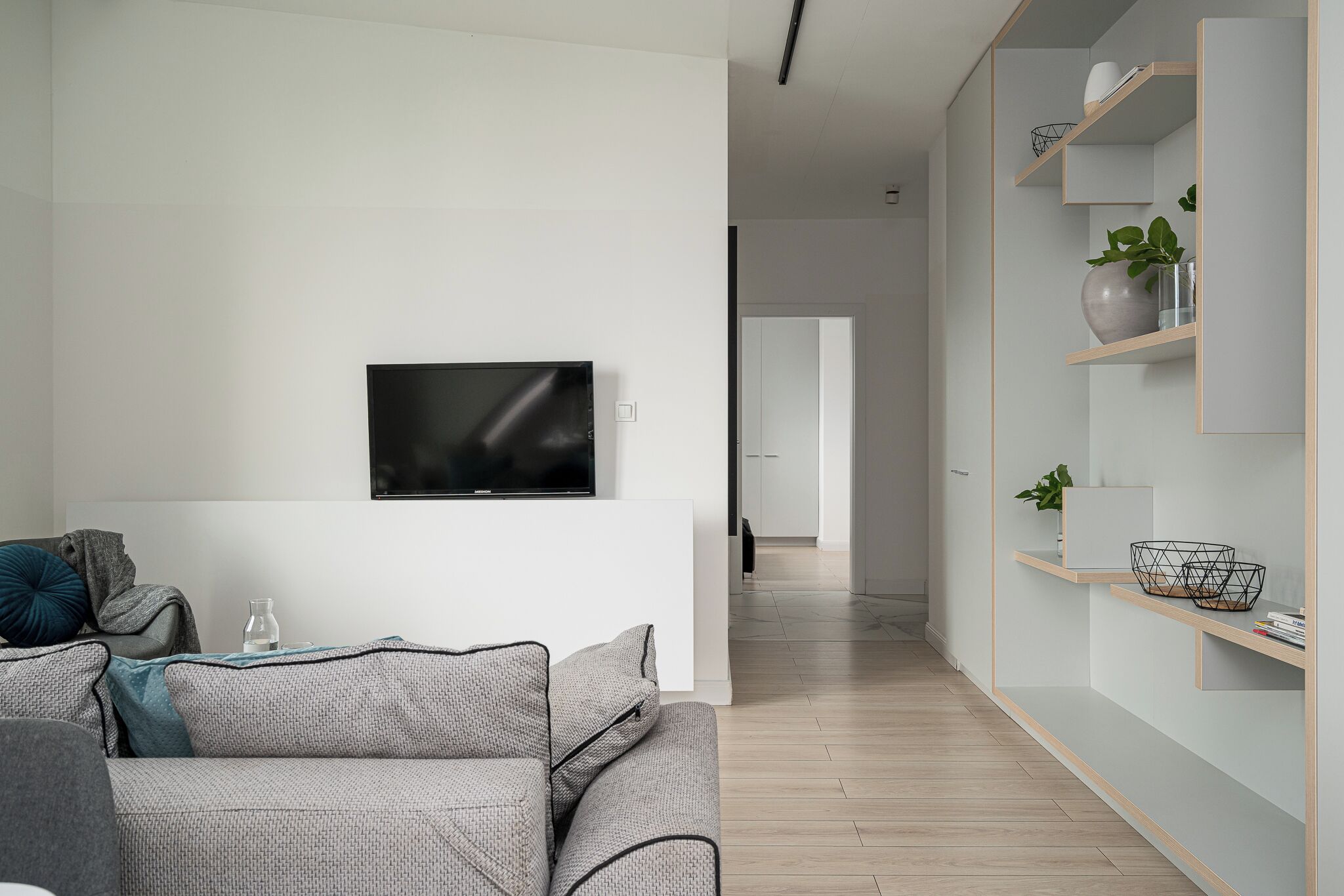Een moderne woonkamer met een flatscreen televisie, een grijze bank en houten schappen met decoratieve objecten.