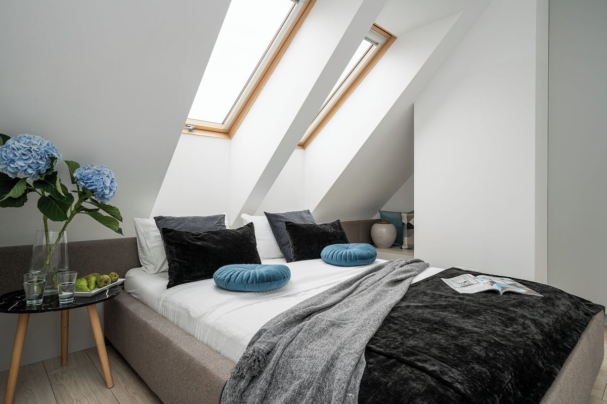 Une chambre moderne avec un plafond en pente, des fenêtres de toit, un lit recouvert d’une couverture grise, deux oreillers bleus et une petite table sur laquelle se trouvent des verres et une plante.