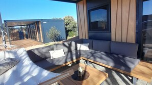 A modern outdoor seating area with a sofa and a wooden table, a large window, and a view of a building and trees. - Gabrijela House (Pirovac)