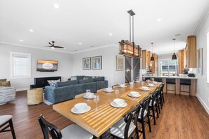 Una cocina moderna con armarios de madera, un comedor con una mesa grande dispuesta para seis personas, y una sala de estar con un sofá y un televisor.