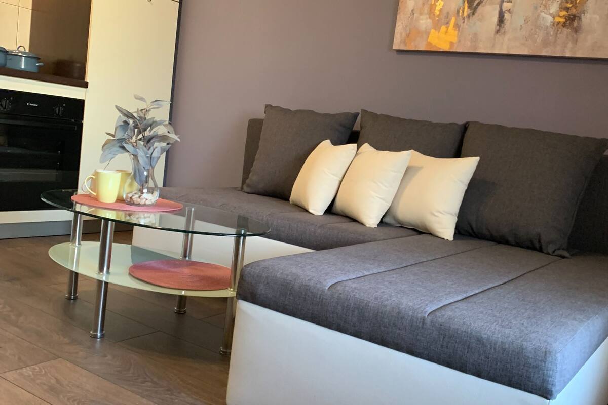 A modern living room with a grey sofa, a glass coffee table, and a kitchen area in the background.