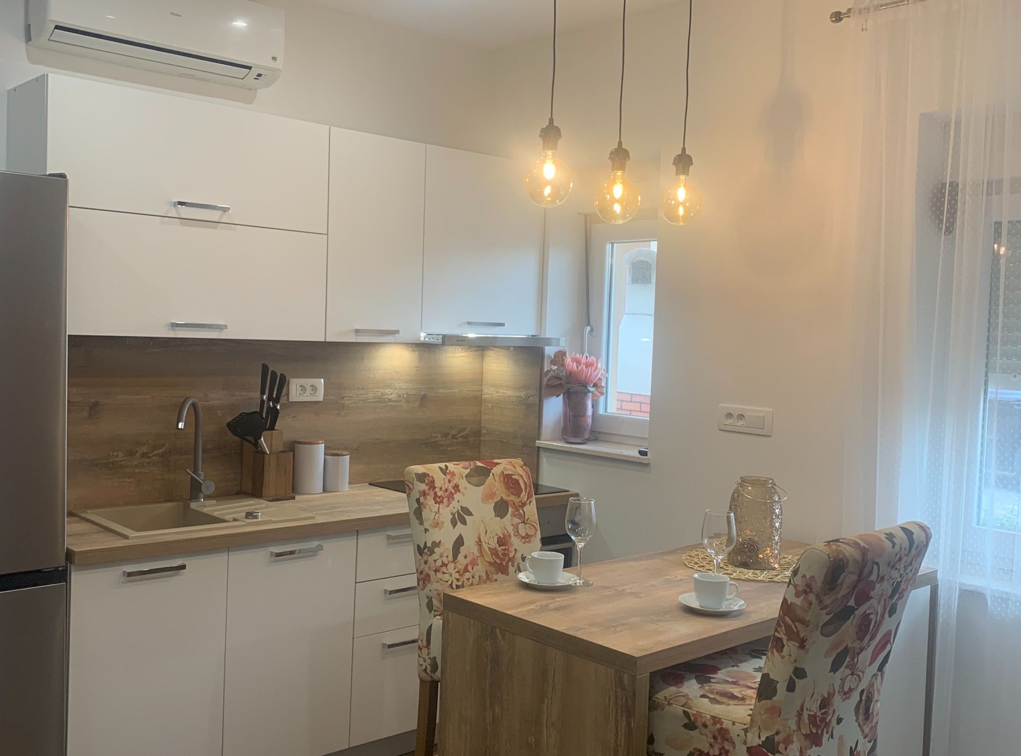 A modern kitchen with white cabinets, a wooden countertop, and a dining table with floral chairs. There are hanging light bulbs above the table.
