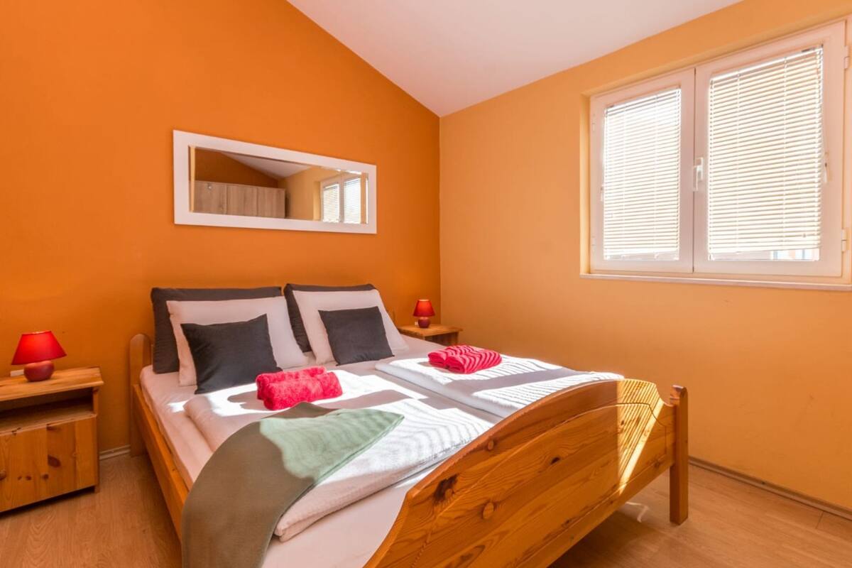 A bedroom with a wooden bed, two bedside tables, a mirror, and a window with blinds.
