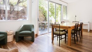 A spacious room with a wooden dining table and chairs, a green armchair, and a large sliding glass door leading to an outdoor area with a brick wall and greenery.