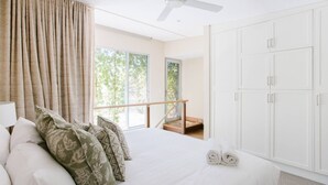 A bedroom with a large bed, a ceiling fan, a balcony with a wooden railing, and a view of trees through the window.