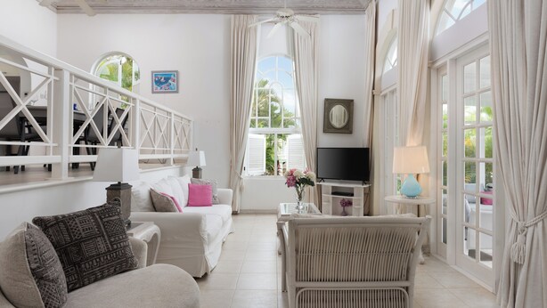 Uma sala de estar ampla com um sofá branco, uma poltrona de vime, uma televisão e uma grande janela com cortinas.