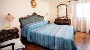 Un dormitorio con cabecera capitonada, cama con manta a rayas, mesita de noche con lámpara, cómoda, espejo y ventana con cortinas.