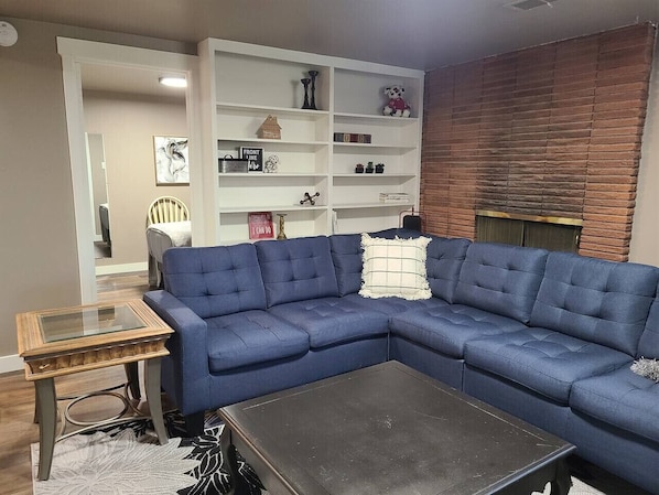 A living room with a blue sectional sofa, a glass-top coffee table, and a white shelving unit with decorative items.
