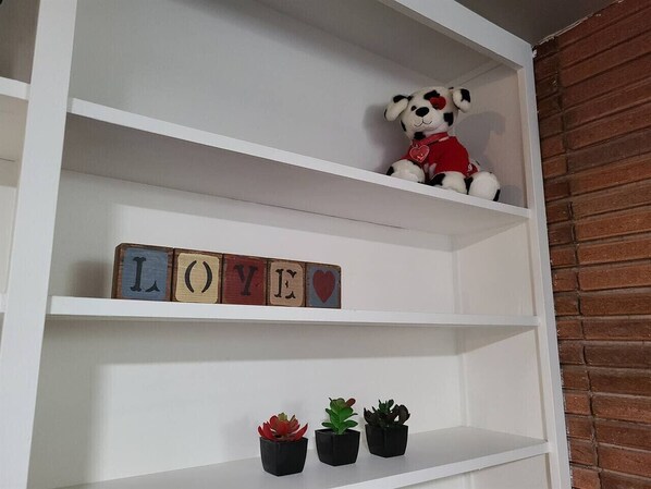 Una estantería blanca con un osito de peluche, bloques de madera que deletrean 'LOVE' y tres macetas con plantas.