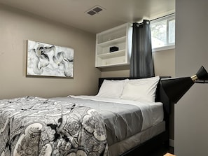 A bedroom with a bed, a wall-mounted shelf, a painting, and a window with curtains.