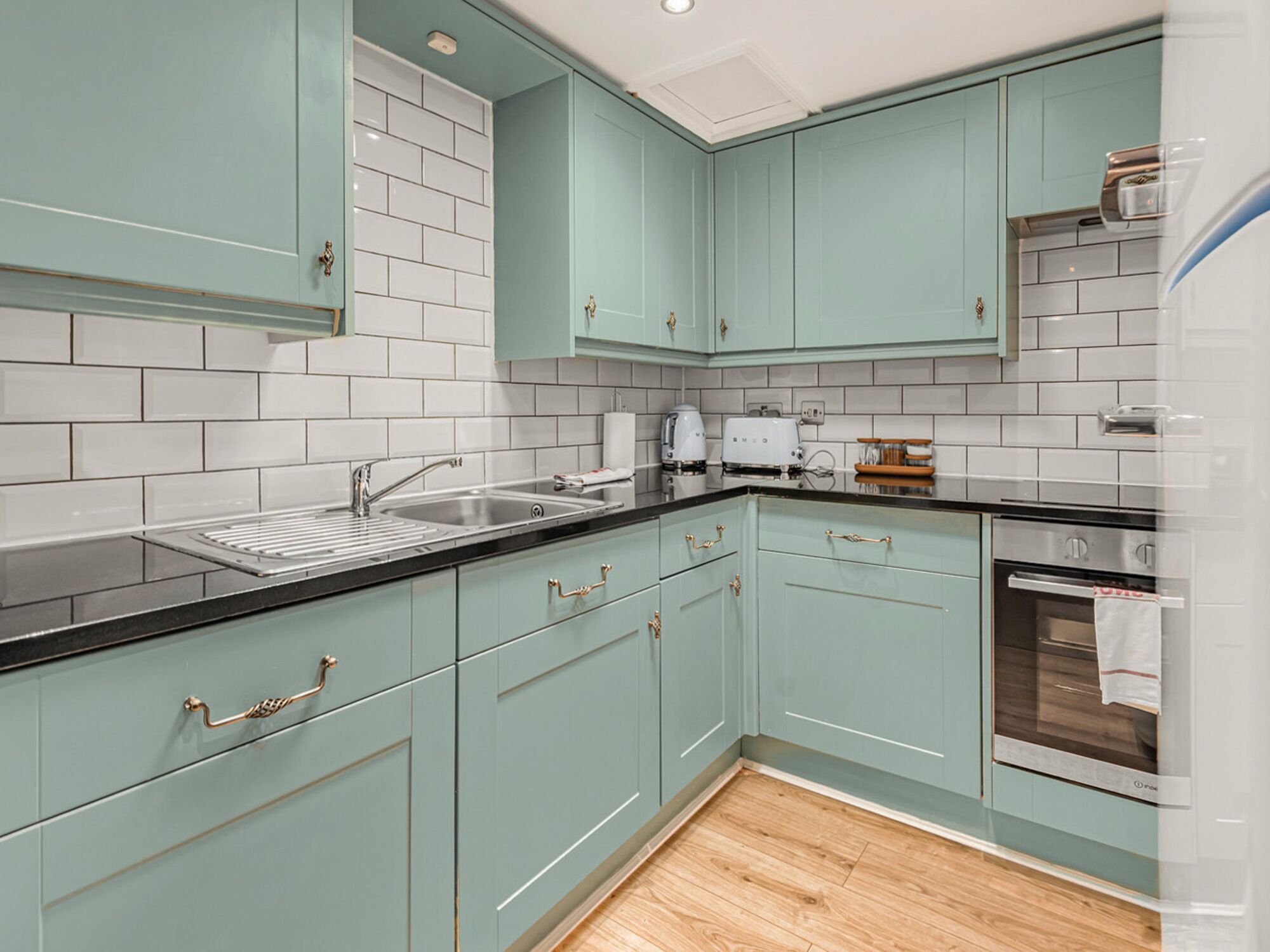 A modern kitchen with teal cabinets, white subway tiles, a stainless steel sink, and a built-in oven.