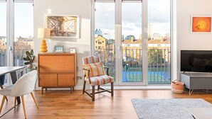 A spacious living room with a large window, a wooden cabinet, a colorful chair, a TV, and a balcony with a view of the city. - Perfectly Located Comfy 1-Bed in London Bridge - Pass the Keys (London)
