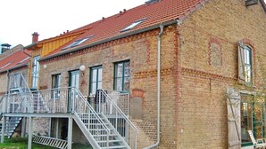 A two-story brick building with a red-tiled roof, a metal staircase leading to a balcony, and a window with a shutter.