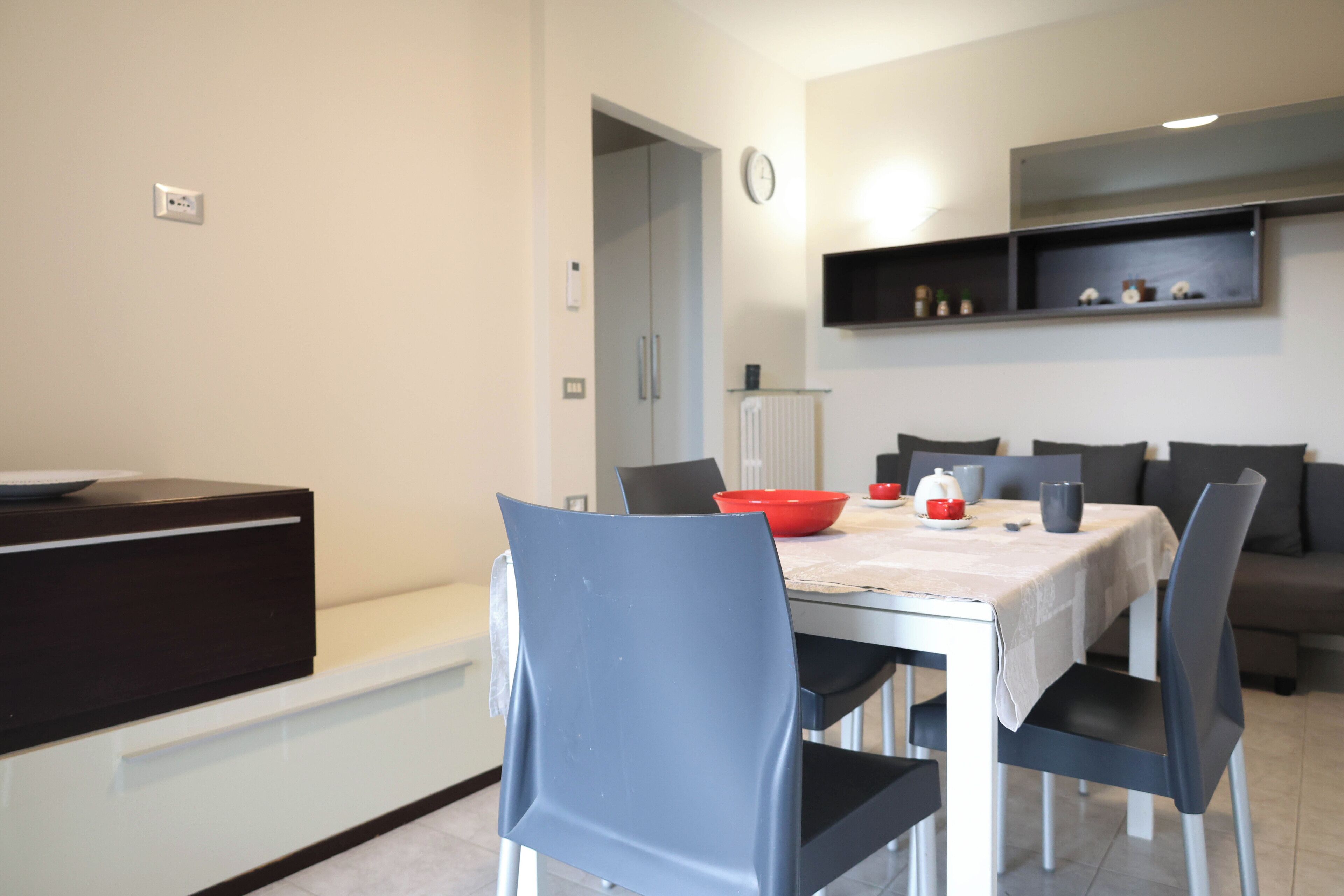 Un espace repas moderne avec une table dressée pour quatre personnes, un meuble de couleur sombre et une étagère murale agrémentée d’objets décoratifs.