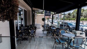 Un caffè all'aperto con tavoli e sedie, un cartello per Le Paradou Café - Restaurant, e vista su auto parcheggiate e edifici.