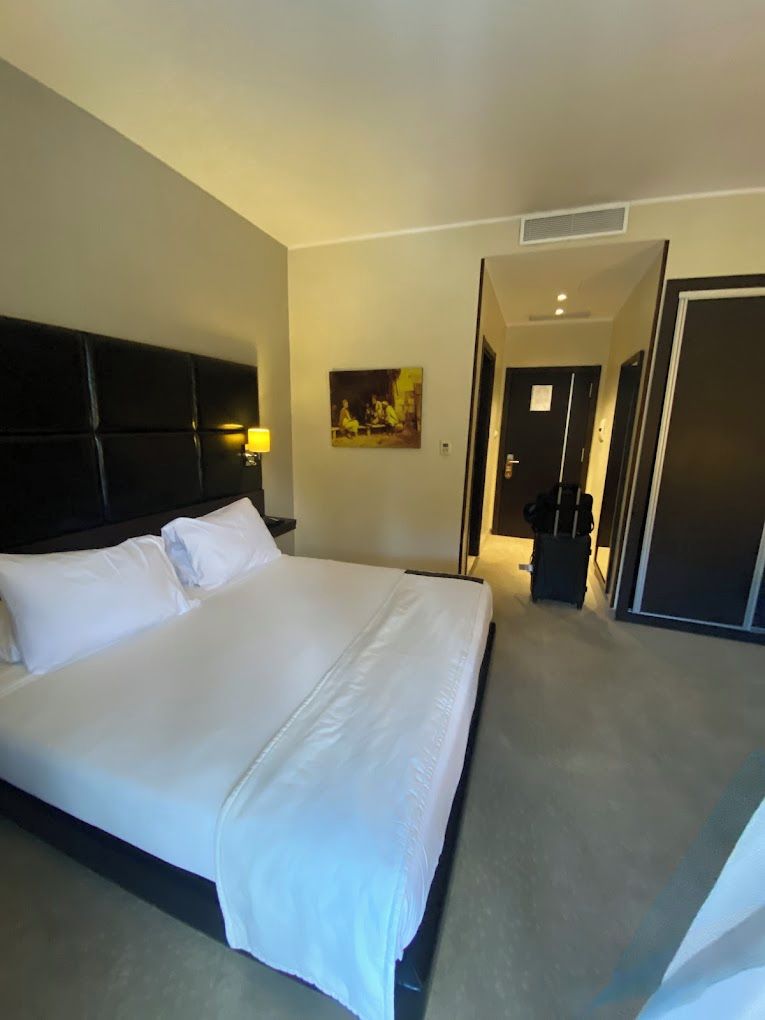 A hotel room with a large bed, a headboard, a painting on the wall, and a door leading to another room.