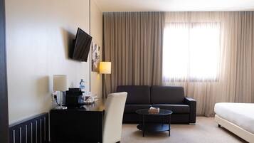 A hotel room with a bed, a sofa, a coffee table, a desk with a lamp, and a TV mounted on the wall.
