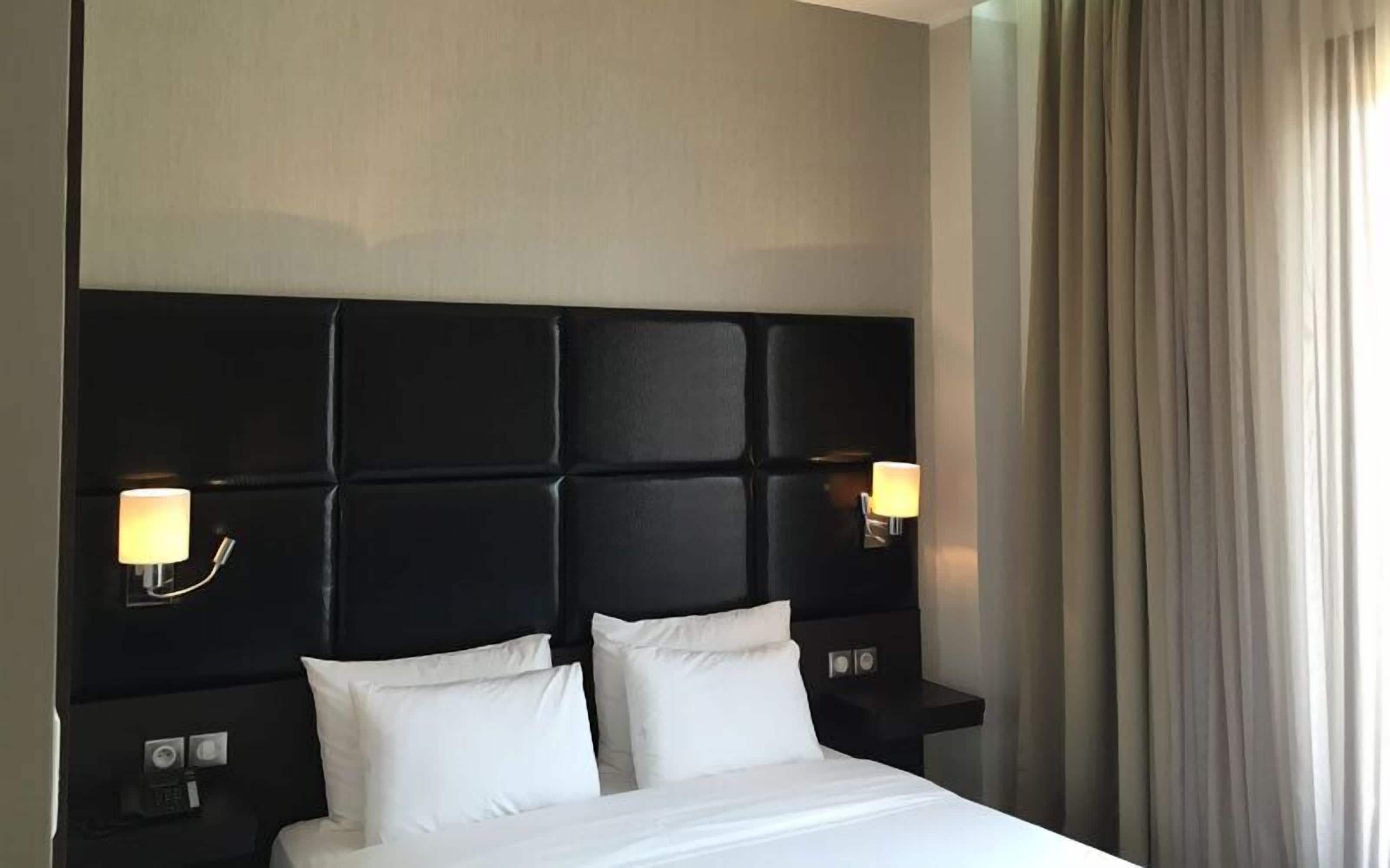 Habitación de hotel con cabecera negra, dos lámparas de noche, cama con ropa de cama blanca y una ventana con cortinas.