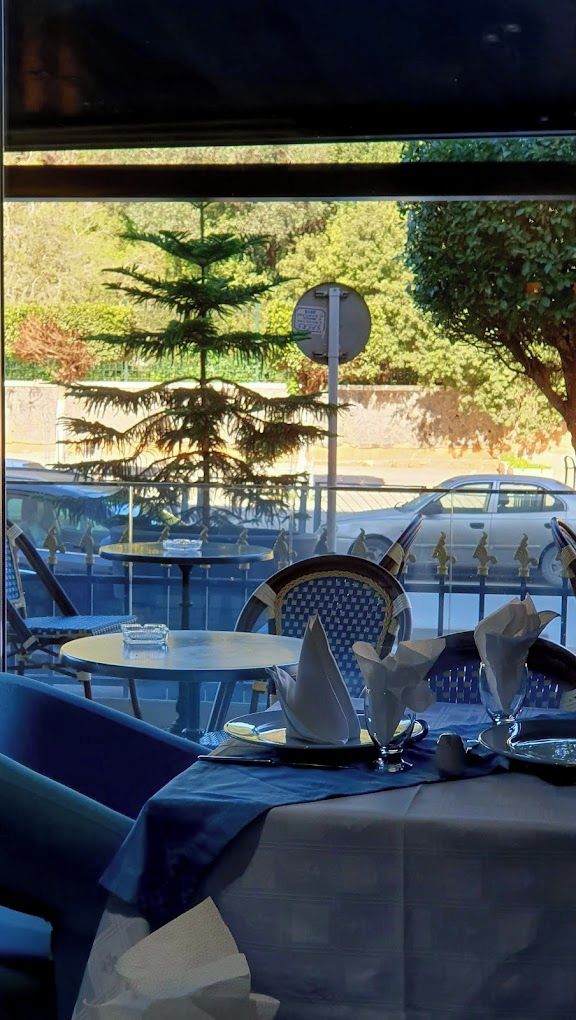 A restaurant with outdoor seating, a table set with a white tablecloth and silverware, and a view of trees and a street with cars.
