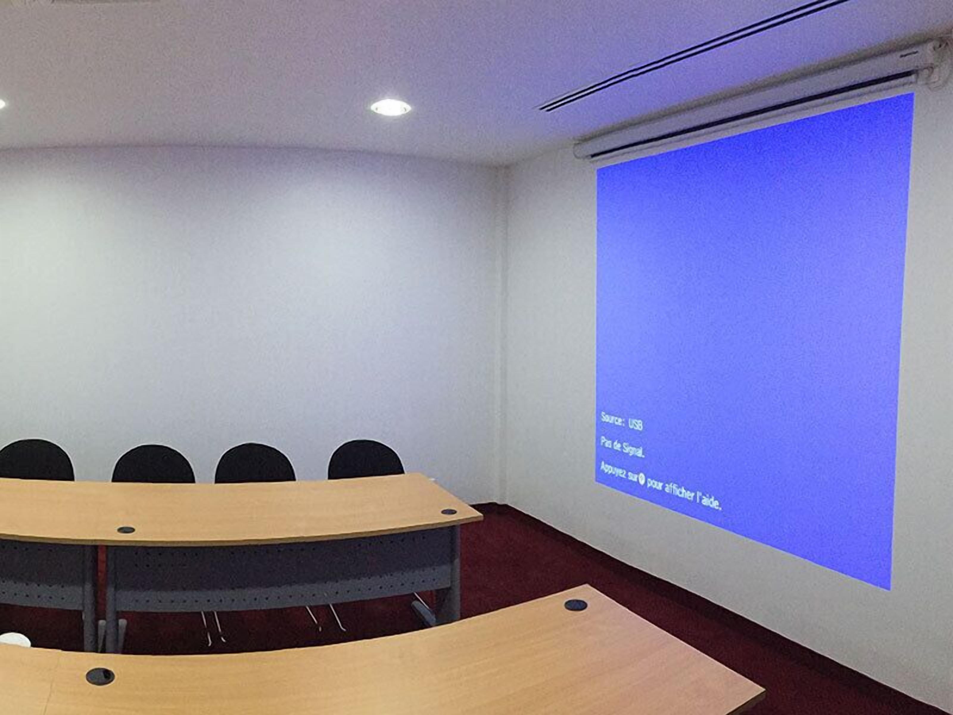 A conference room with a large screen displaying text, a wooden table, and several chairs.