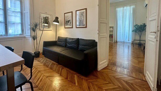 A modern living room with a black leather sofa, wooden herringbone flooring, a dining table with chairs, and a floor lamp. - Casa Milano Classic Elegance with Balcony (Milan)