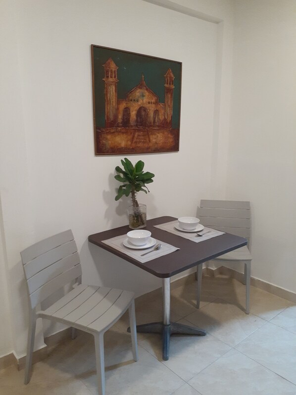 A dining area with a table set for two, two white chairs, a vase with a plant, and a painting of a building on the wall. - Charly's Place  (San Salvador)