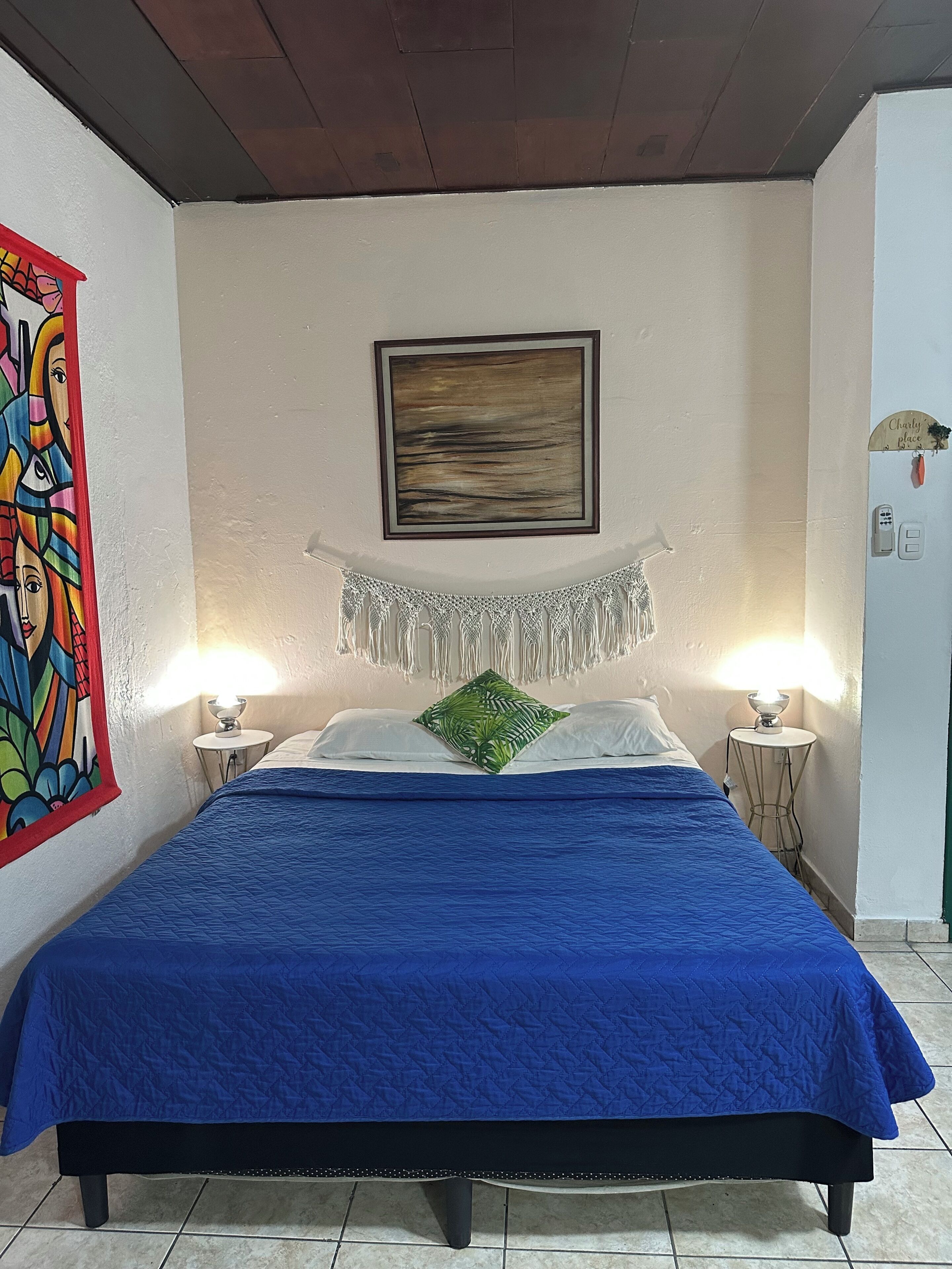 Un lit avec une couette bleue, un oreiller vert et un tableau mural. Il y a un tableau encadré au-dessus du lit et une petite table avec une lampe à côté.