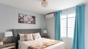 Un dormitorio con una cama, dos mesitas de noche, aire acondicionado de pared y una ventana con cortinas.