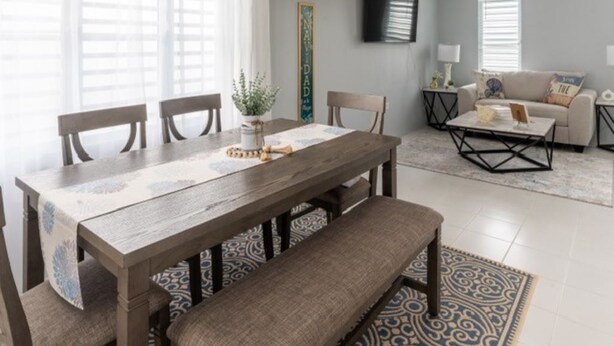 Un comedor con una mesa y sillas de madera, un banco y un tapete estampado. En el fondo, hay un sofá y una mesa de centro.