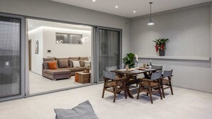 A modern living room with a sofa, a dining table with chairs, and a large window. - Zoe Luxury Seafront Villa with Heatable Pool, Villa (Heraklion)
