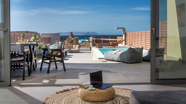 A balcony with a pool, seating, and a laptop on a woven pouf. - Zoe Luxury Seafront Villa with Heatable Pool, Villa (Heraklion)