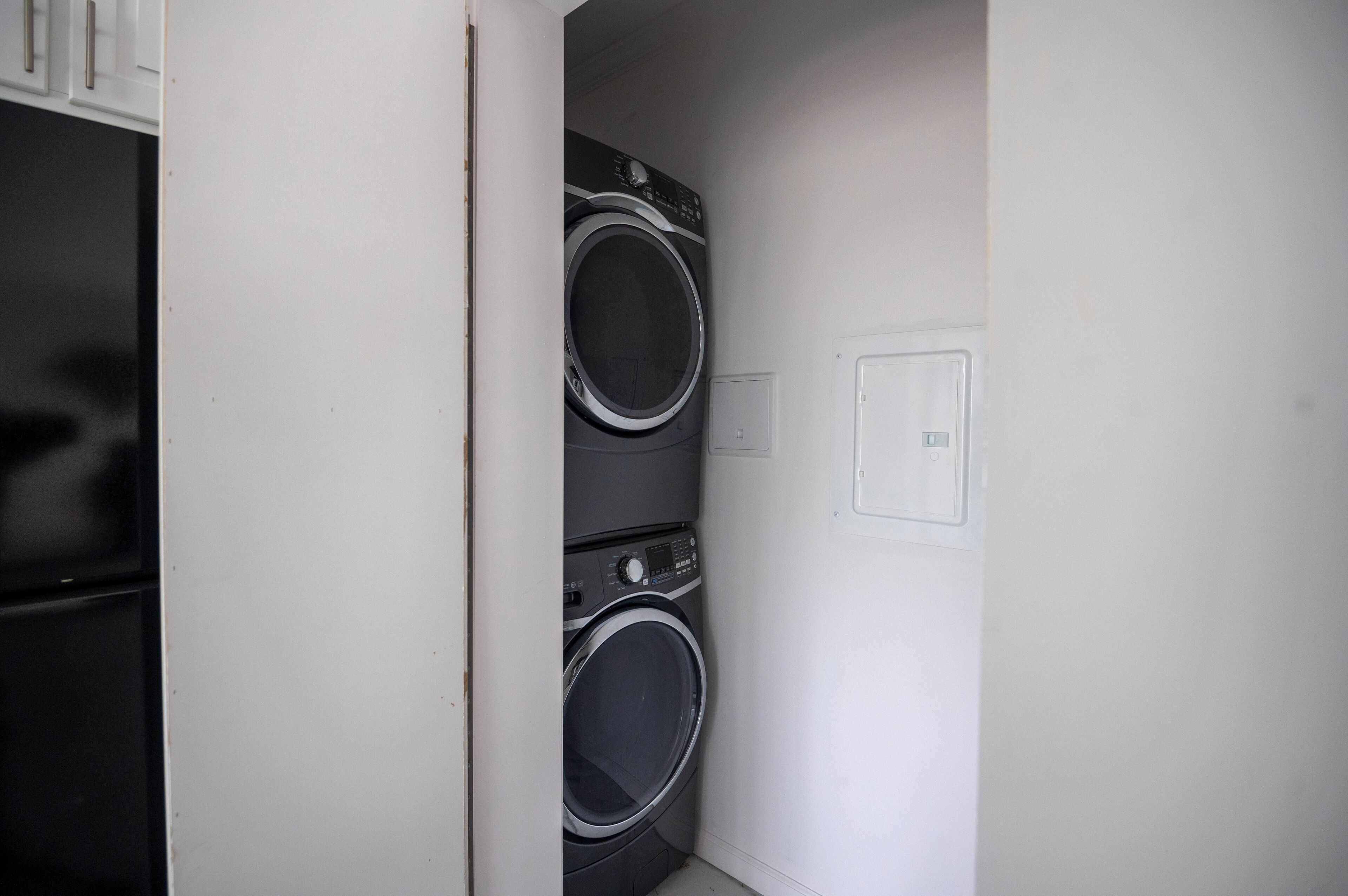 Uma lavanderia com duas máquinas de lavar frontais empilhadas e uma porta que dá para outro espaço.
