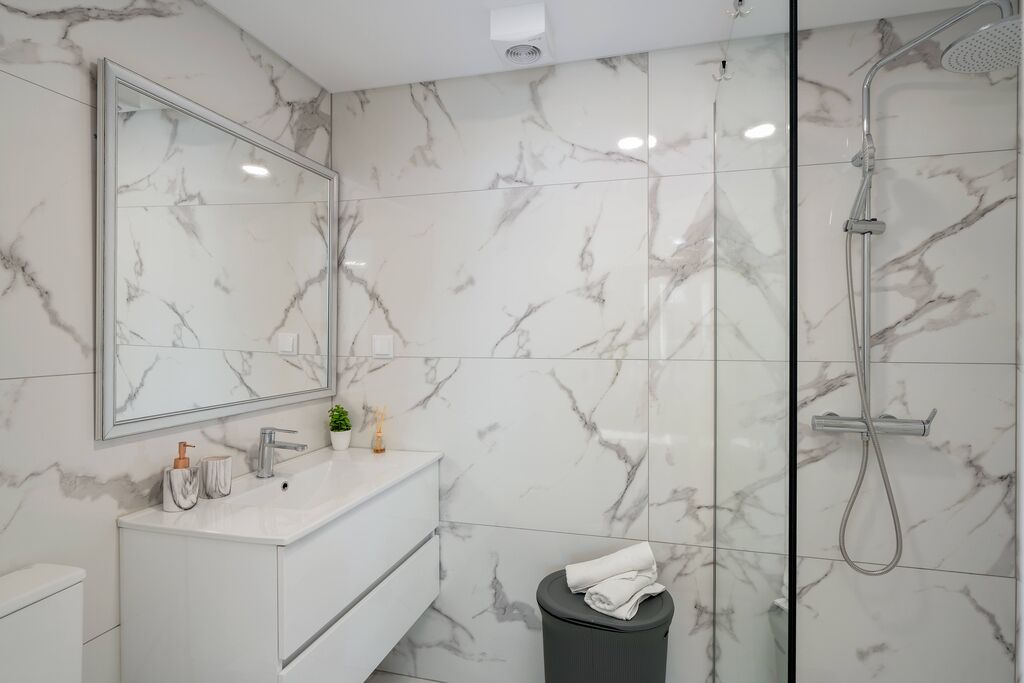 Et moderne badeværelse med en marmorbeklædt bagvæg, et stort spejl, et hvidt toiletbord og en bruser med håndbruser.