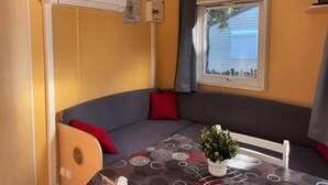 A compact room with a built-in sofa, a small table with a potted plant, and a window with a view of a house. - MOBILE-HOME 1 BEDROOM WITH AIR-CONDITIONING SOUTH ARDÈCHE (07) ORGNAC-L'AVEN (Orgnac-l'Aven)