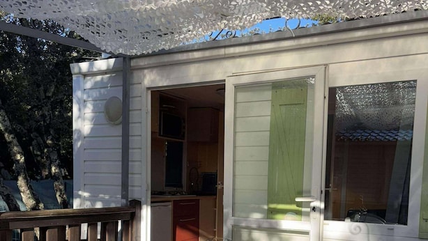 A covered outdoor dining area with a table and chairs, a wooden deck, and a building with a green door and windows. - MOBILE-HOME 1 BEDROOM WITH AIR-CONDITIONING SOUTH ARDÈCHE (07) ORGNAC-L'AVEN (Orgnac-l'Aven)