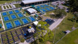 Luchtbeeld van een sportcomplex met meerdere tennisbanen, een overdekt zitgedeelte en een parkeerplaats.