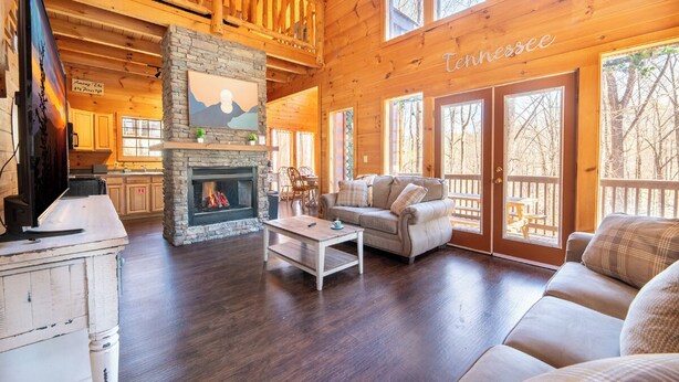 A cozy cabin interior with a stone fireplace, wooden floors, a sofa, and large windows. - *Spacious 4BD Lodge w/ POOL! Hot Tub + GAMES!* (Sevierville)