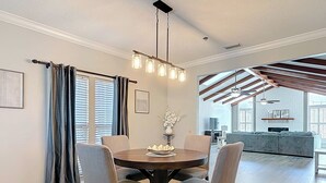 A dining area with a round table, four chairs, and a chandelier. There is a living area with a sofa and a TV in the background.