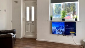 A room with a white door, a window with a view of trees and a car, and a flat-screen TV mounted on the wall displaying a photo gallery. - Cosy Contractors Retreat in Batley - Sleeps 3 (Batley)