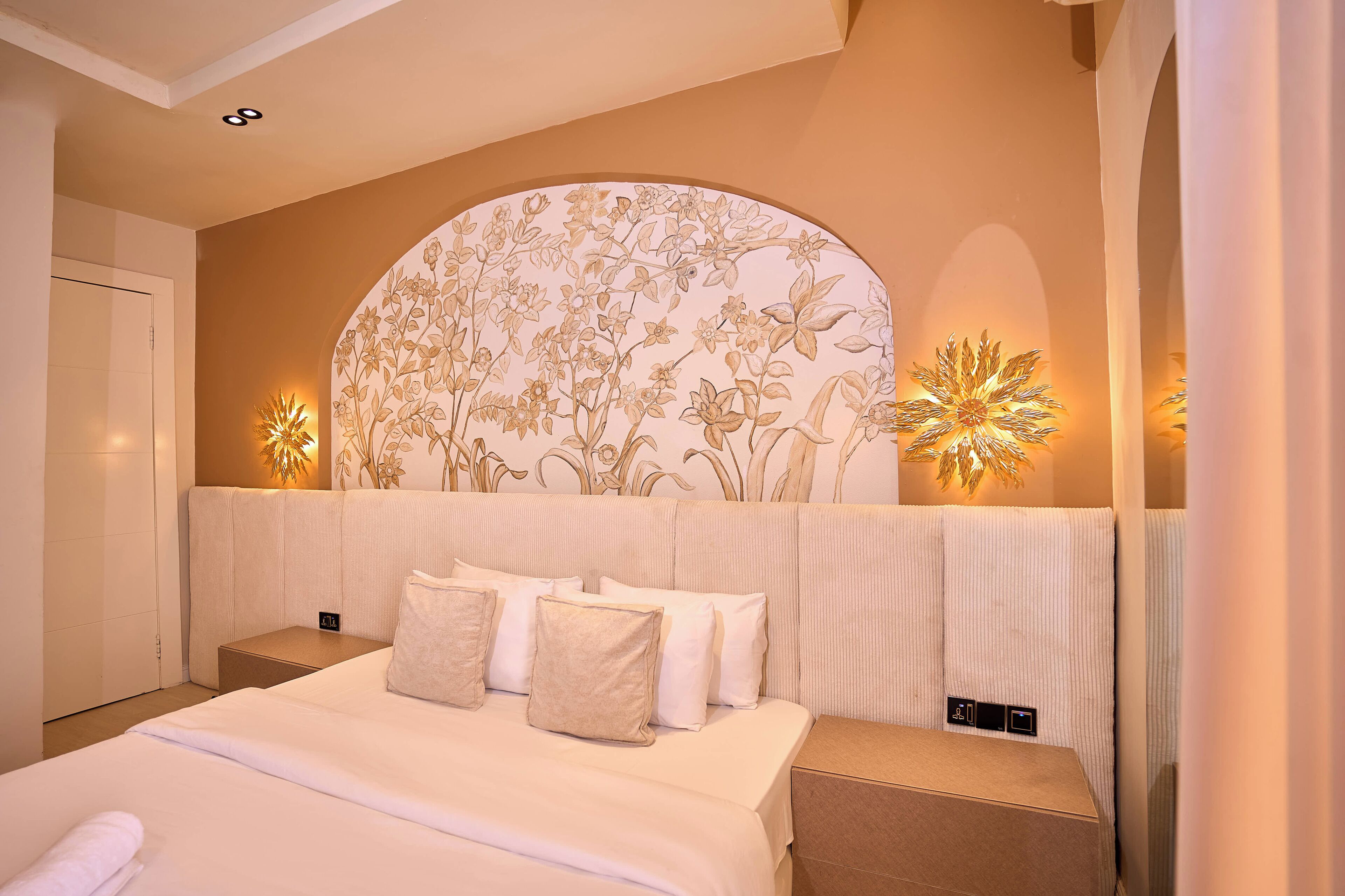 A hotel room with a large bed, a curved headboard with floral patterns, bedside lamps, and a control panel on the nightstand.