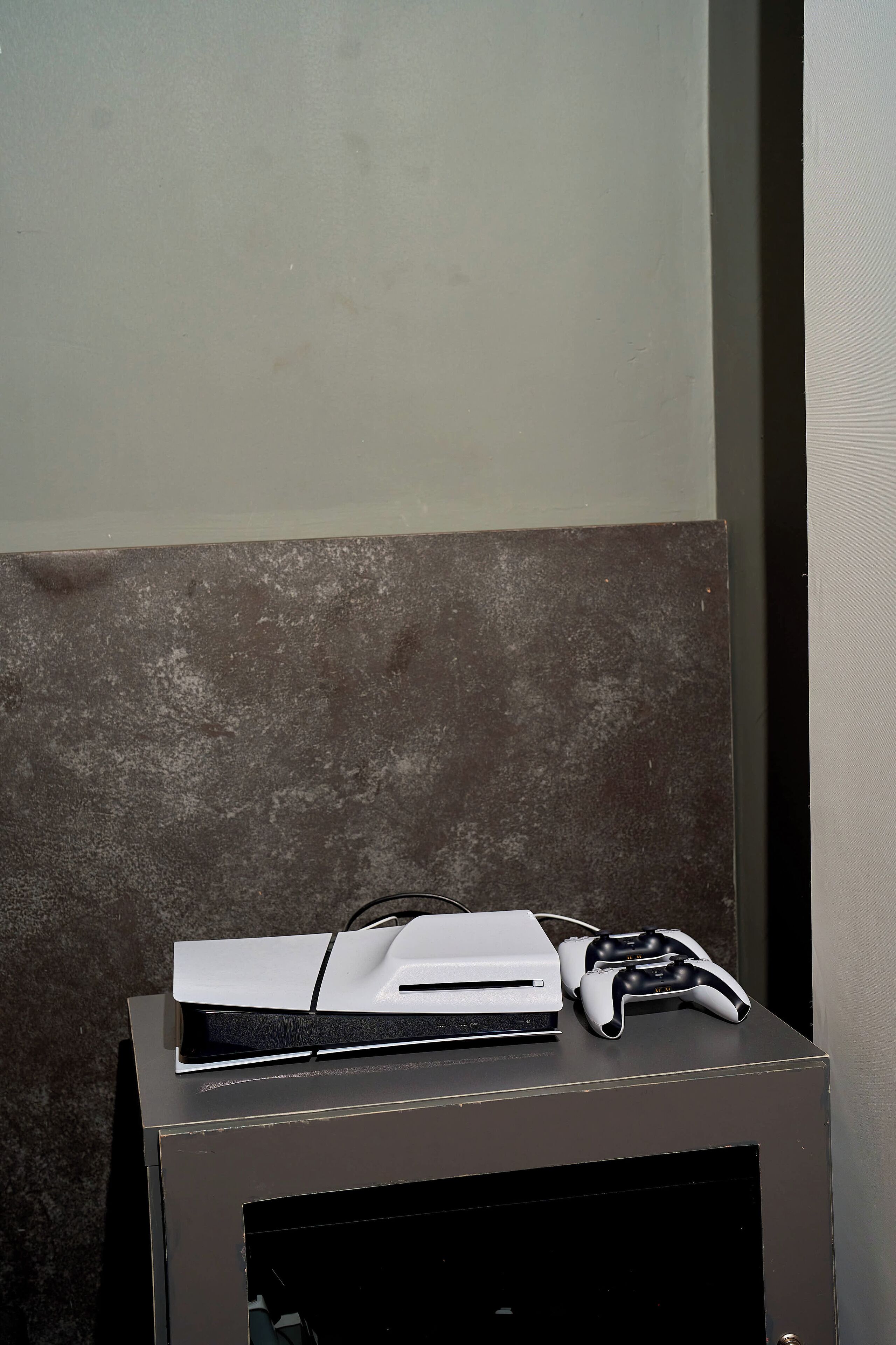 A gaming console and controller on a dark-colored cabinet against a textured wall.