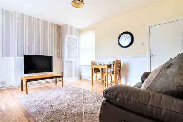 A living room with a flat-screen TV on a wooden stand, a brown leather sofa, a patterned rug, a dining table with chairs, and a wall clock. - Travellers Economic Stays in Orrell Park (Liverpool)