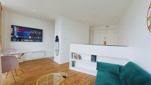 Een moderne woonkamer met een flatscreentelevisie, een glazen salontafel, een witte bank met blauwgroene kussens en een witte boekenkast.