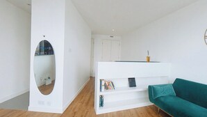 Een moderne, minimalistische woonruimte met een witte muur, een ronde spiegel, een blauwgroene bank en een witte boekenkast met boeken en een afstandsbediening.