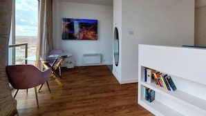 Een moderne woonkamer met een houten vloer, een glazen tafel, een paarse stoel, een witte boekenkast en een groot schilderij aan de muur.