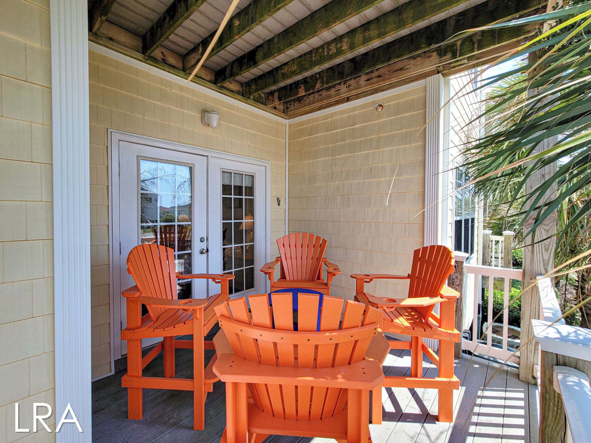 Une terrasse couverte avec des chaises et une table en bois, une porte avec des panneaux de verre et une vue sur l’extérieur.