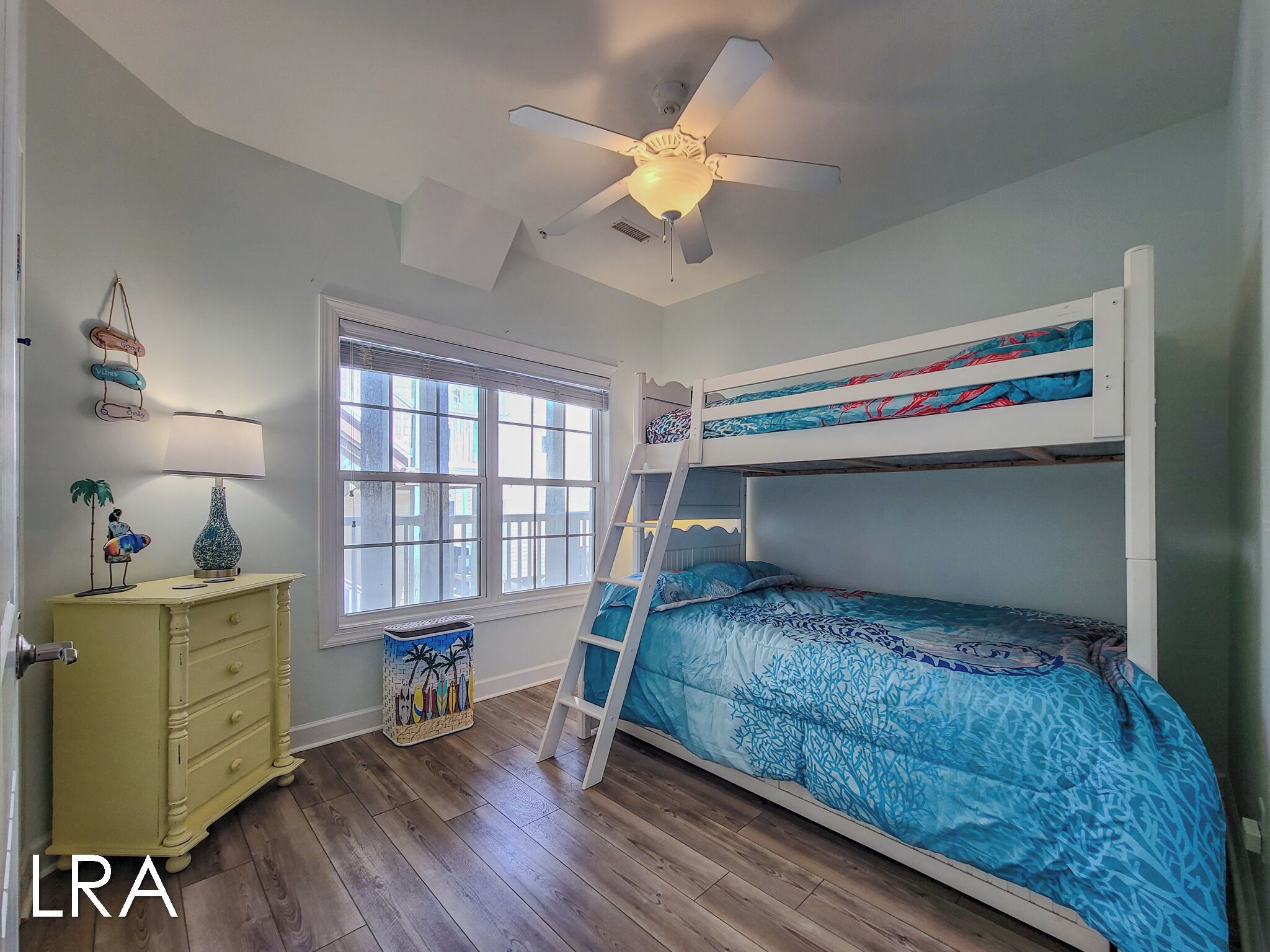 A room with a bunk bed, a desk with a lamp, a window with blinds, and a ceiling fan.