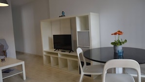A modern living room with a flat-screen TV on a white entertainment unit, a round dining table with two chairs, and a potted plant on the table. - Apartments Taravilla 3D P26, in Bahia Beach (Puerto de Mazarrón)